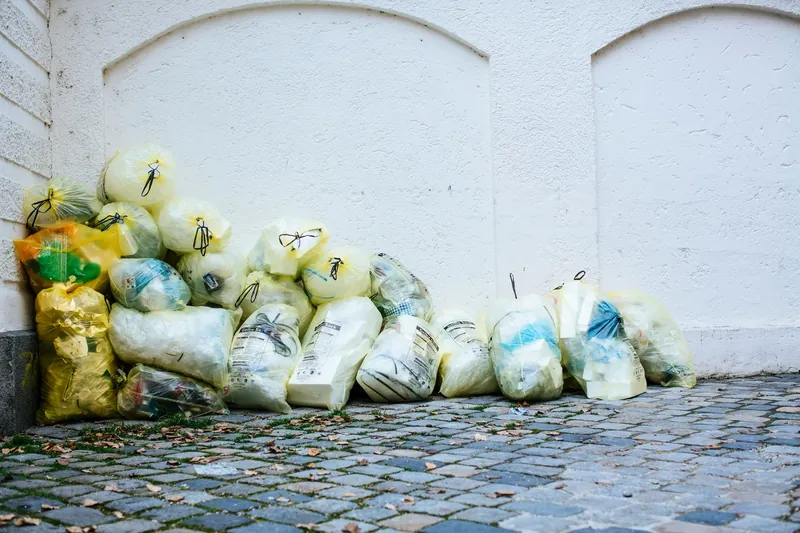 Pile of tied waste bags stacked against a white wall on a paved corner
